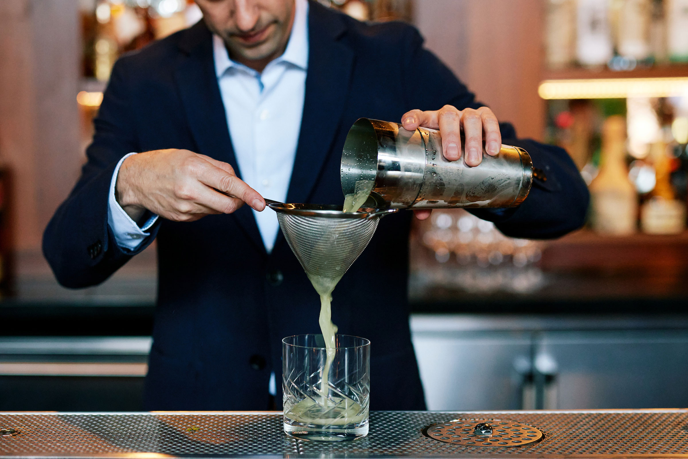 bartender pouring a snap pea cocktail through a sieve and into a crystal tumbler