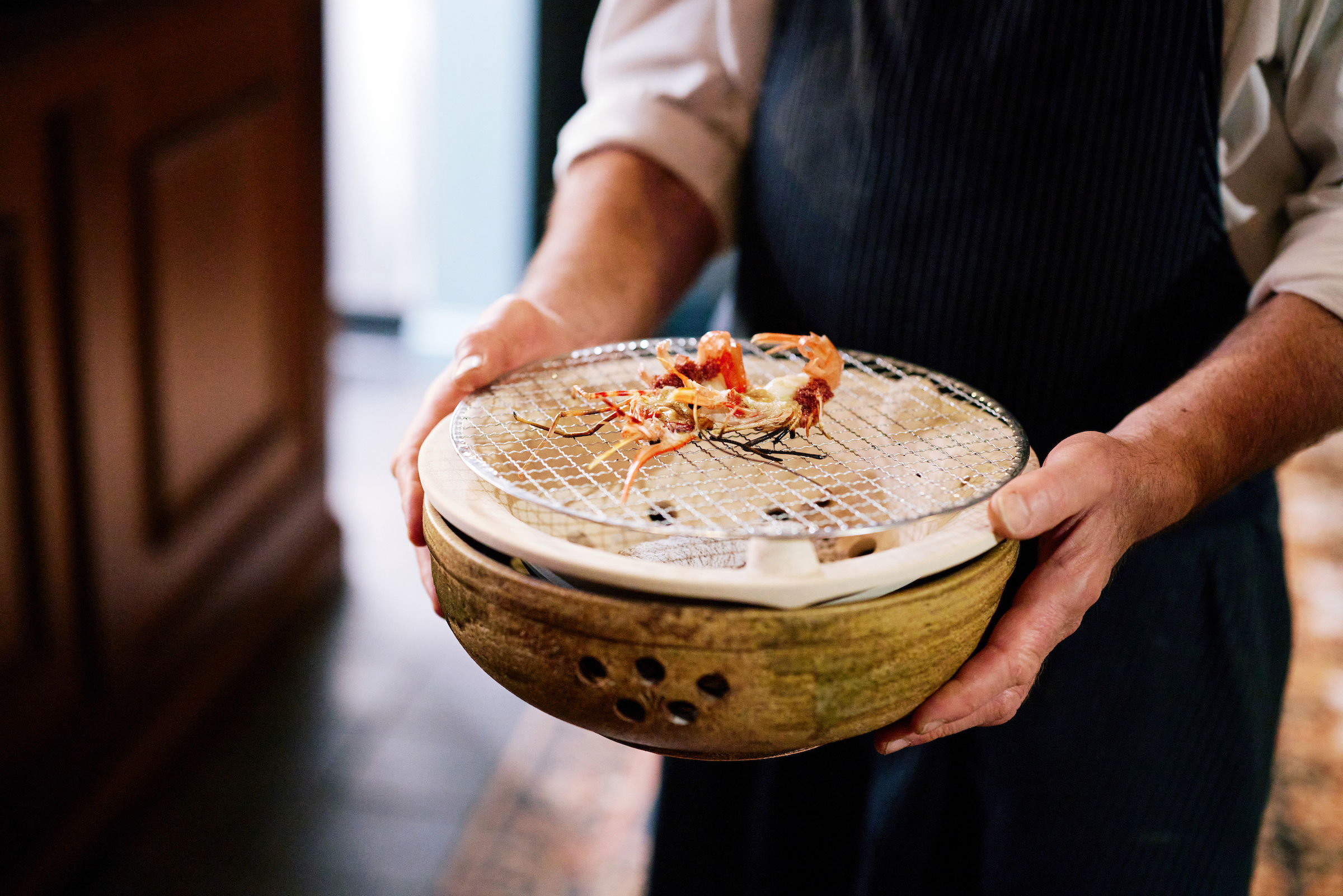 server carrying handmade wooden bowl with metal grill on top for grilling prawns tableside